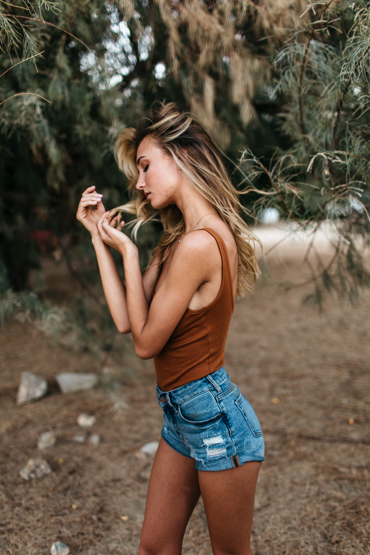 A woman with long blonde hair wearing a brown tank top and denim shorts, standing outdoors near a tree, with her eyes closed and her hands near her face.