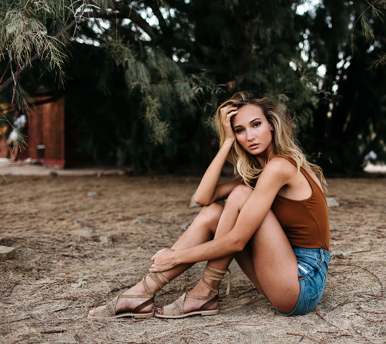 A young woman with blonde hair and a brown tank top sitting on sandy ground among trees, looking at the camera with her hand resting on her head.