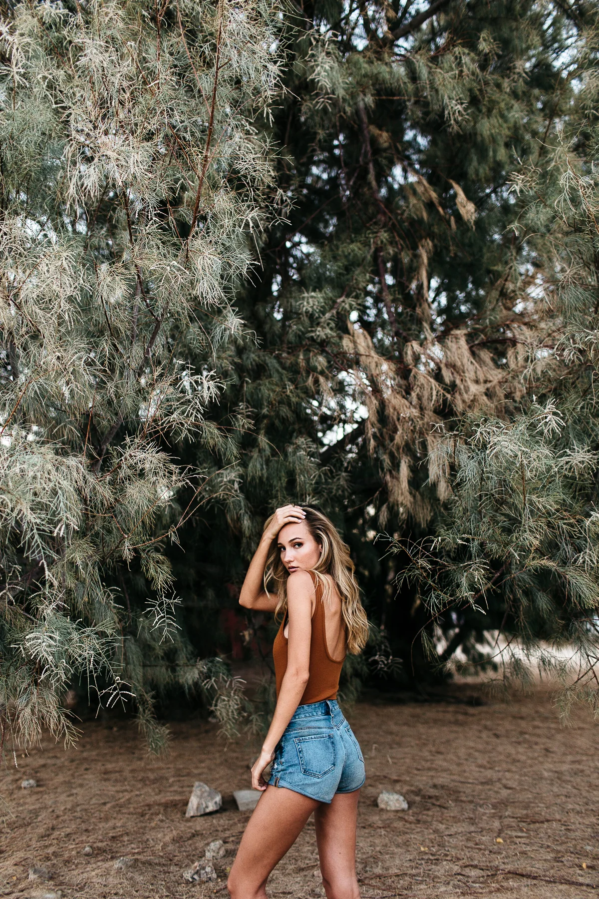 A woman with long blonde hair wearing a brown tank top and denim shorts standing outdoors in front of trees, looking over her shoulder with one hand on her head.