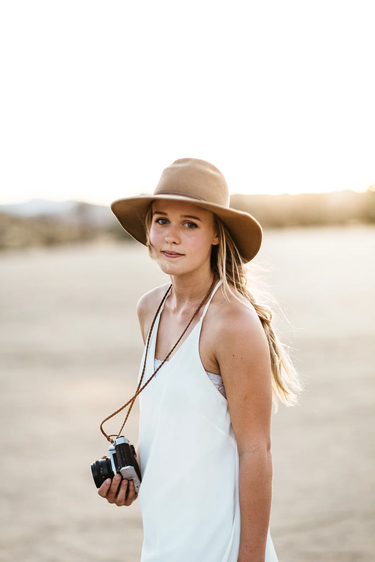A young woman with long blonde hair wearing a beige wide-brimmed hat and white sleeveless dress, holding a vintage camera, outdoors during sunset with blurred landscape in the background.