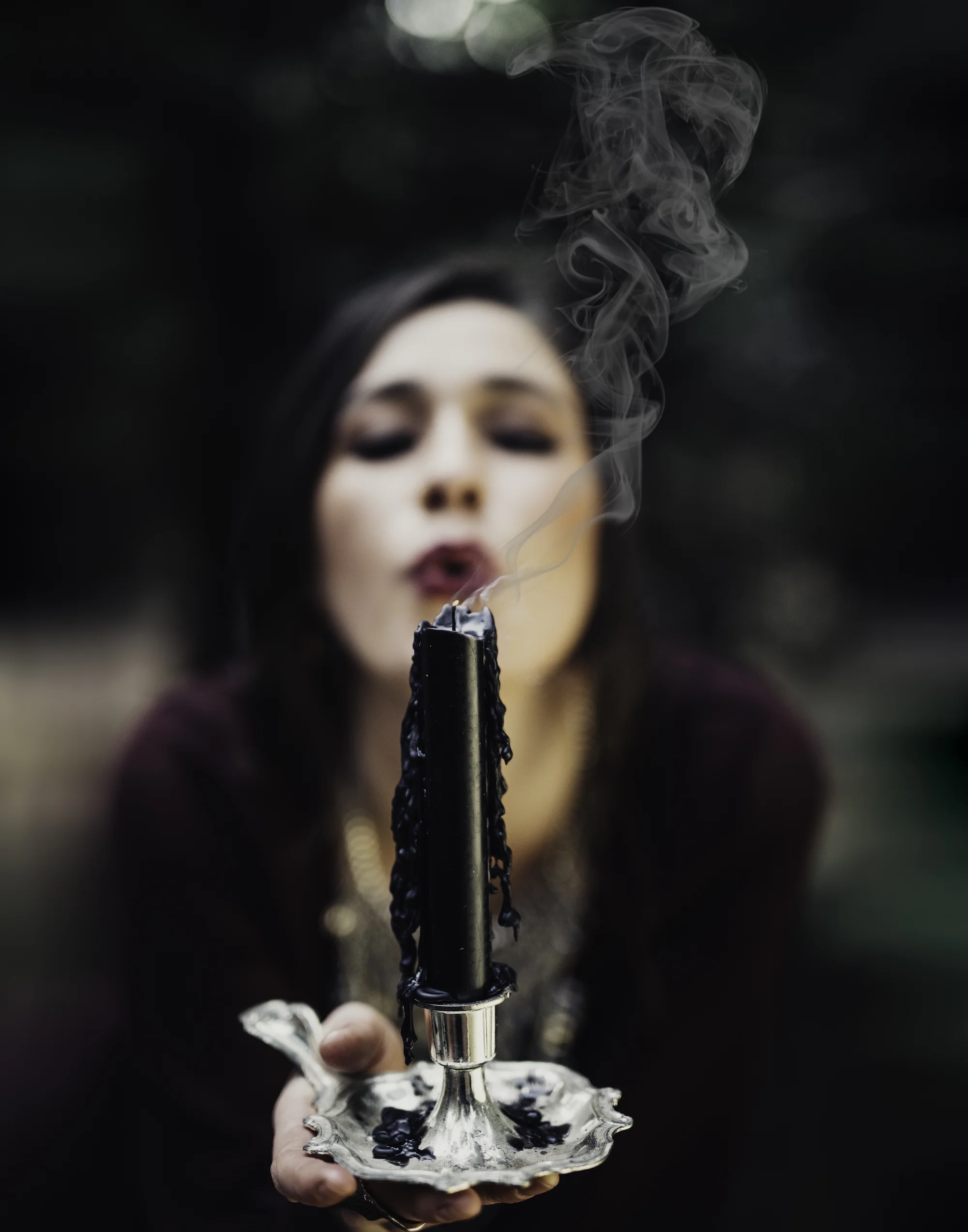 A woman holding a black candle with black wax dripping down, smoke rising from the candle, and a silver candlestick holder, with her face blurred in the background.