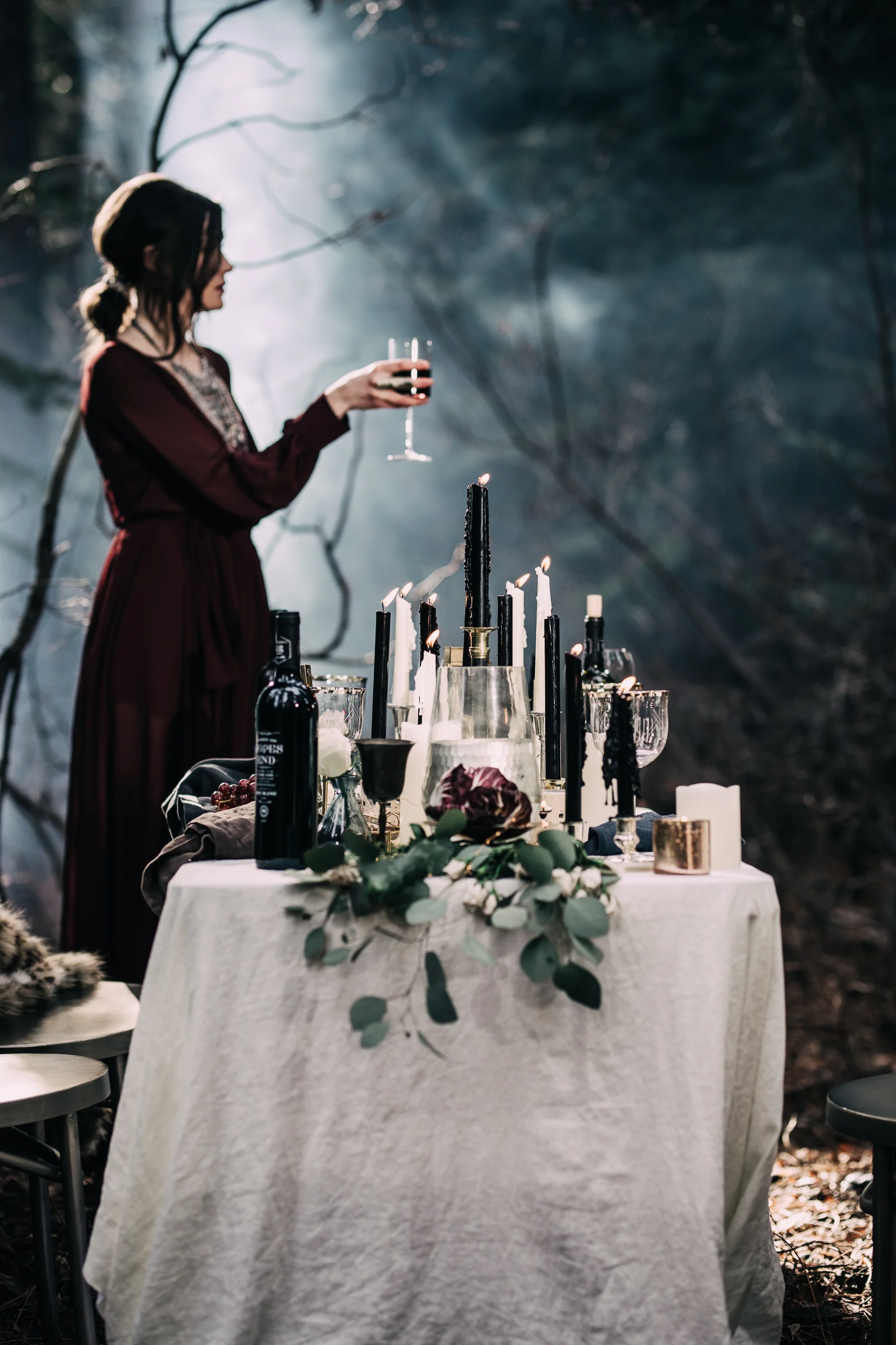 A woman in a burgundy dress standing outdoors at a candlelit table, holding a glass of red wine.