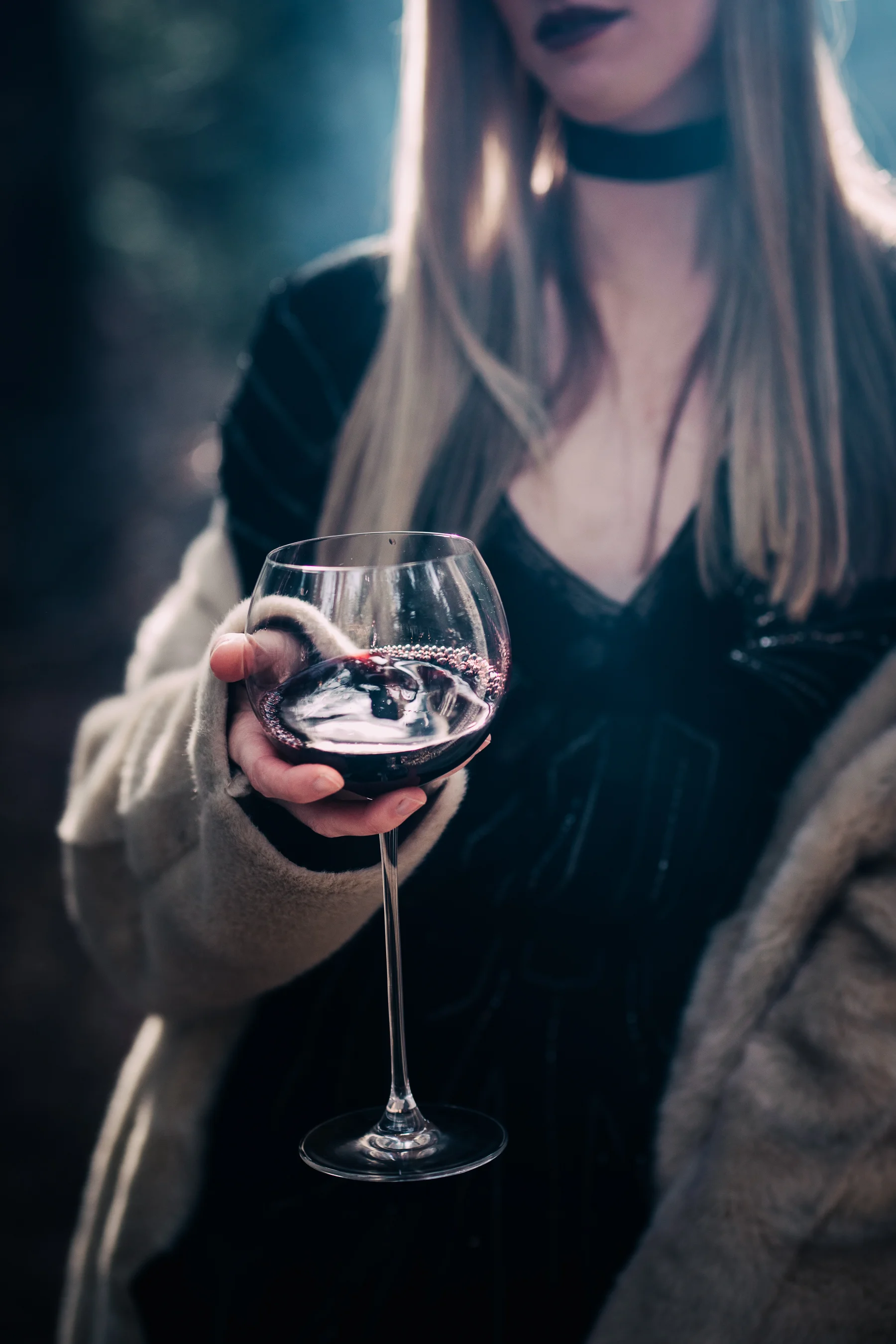 A woman in a black dress and beige cardigan holding a glass of red wine outdoors during evening.