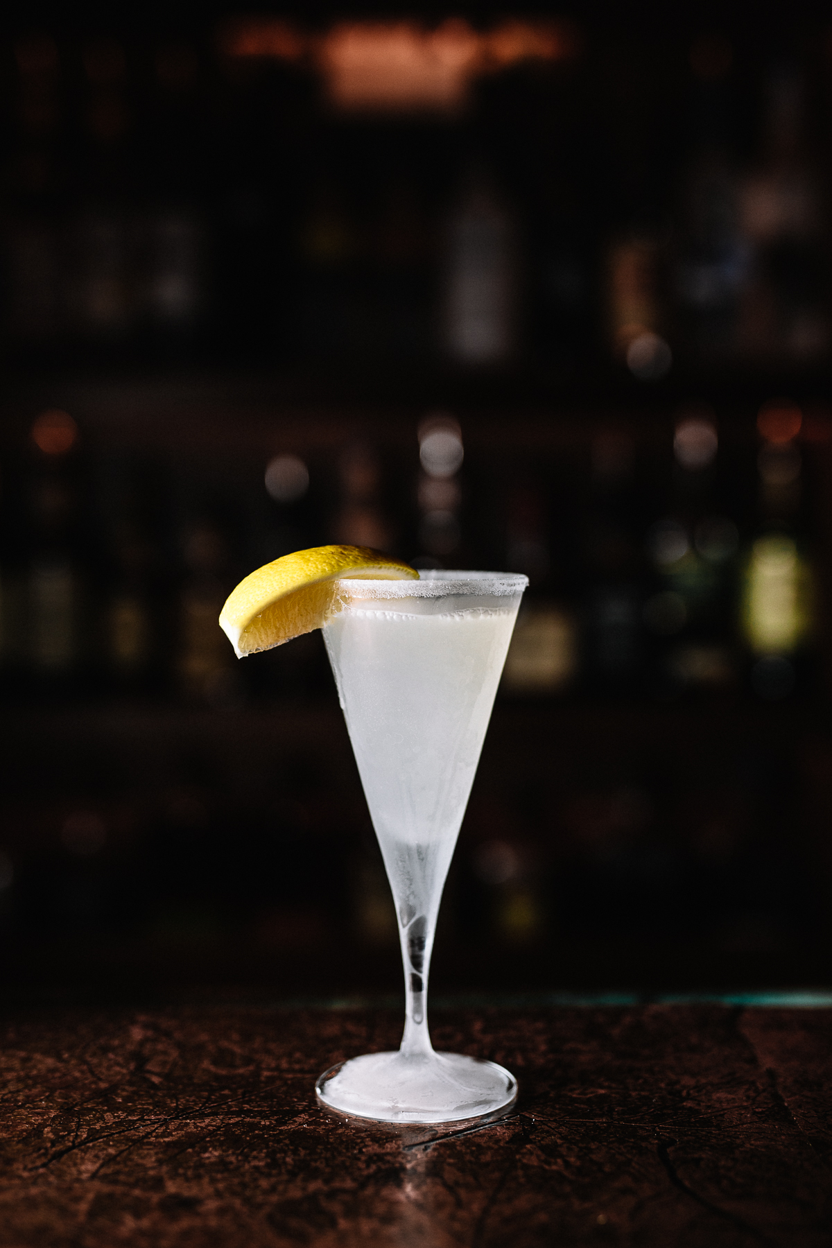A white cocktail with a lemon wedge garnish on the rim, served in a tall, elegant glass on a dark bar counter.