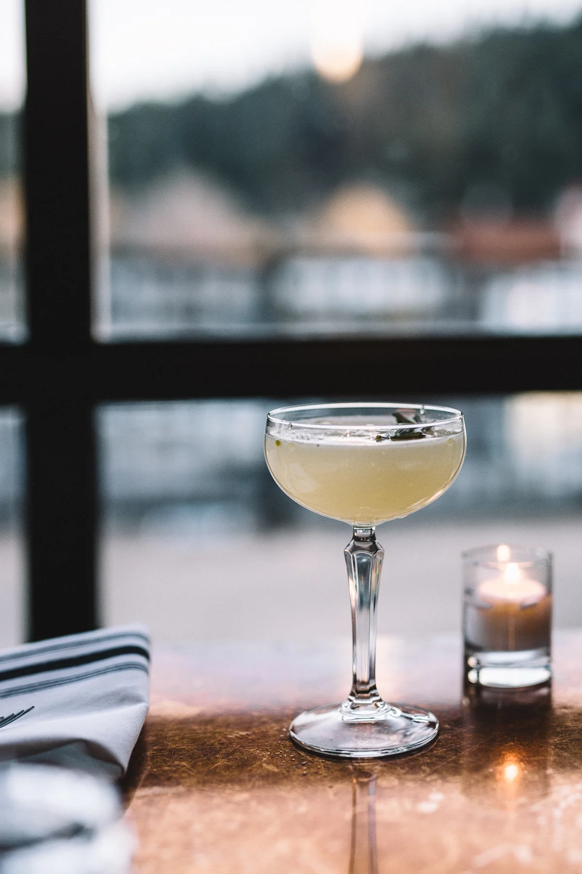 A cocktail in a coupe glass on a wooden table with a lit candle in a glass holder and a napkin with black stripes, with a blurry window in the background.