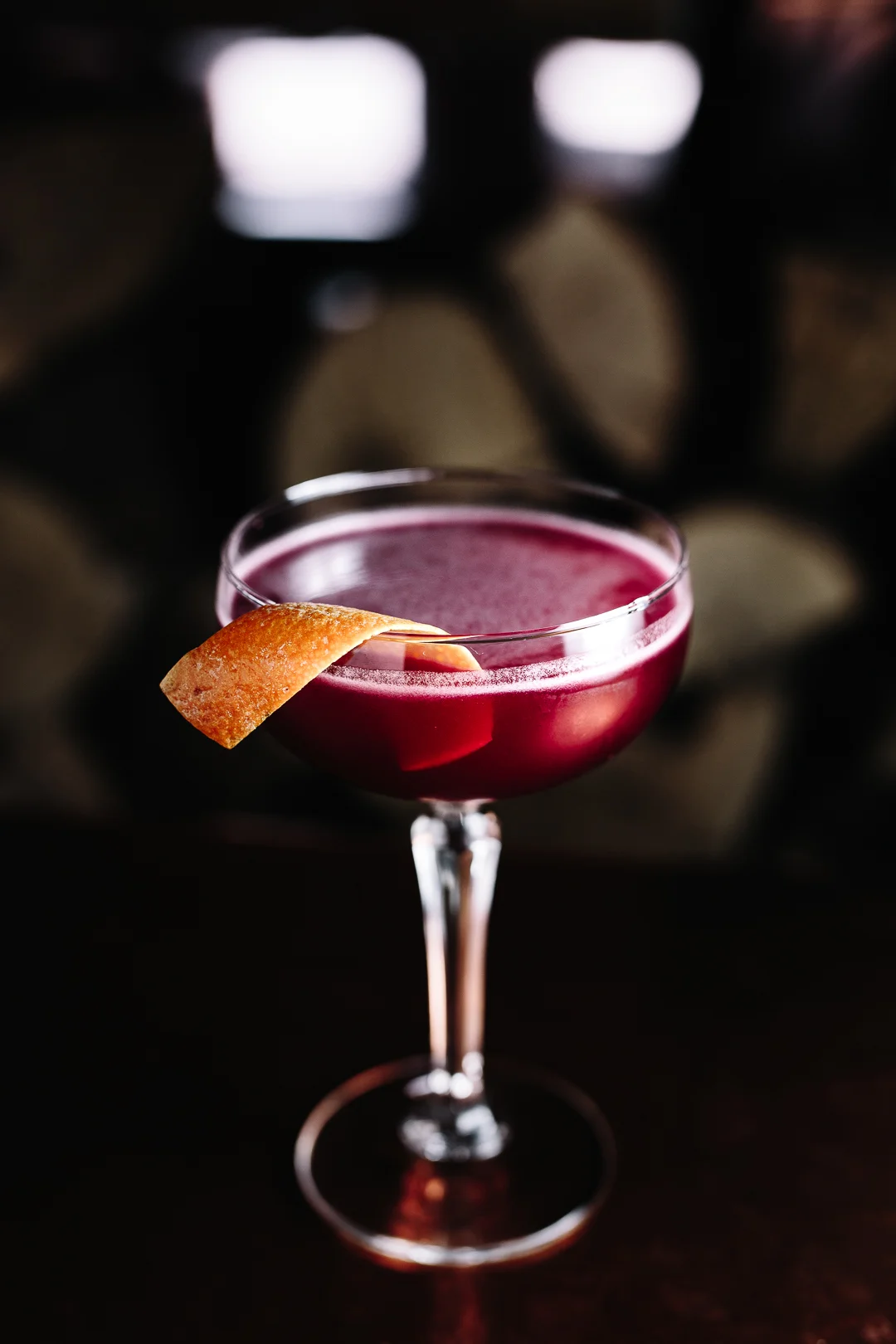A cocktail in a coupe glass with a lemon twist garnish, set against a dark background with blurred barrels.