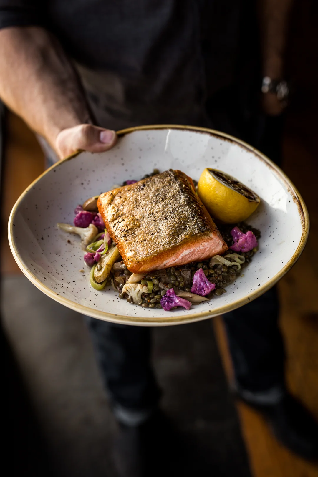 A hand holds a ceramic bowl with a grilled salmon fillet, lemon half, lentils, and purple pickled vegetables.