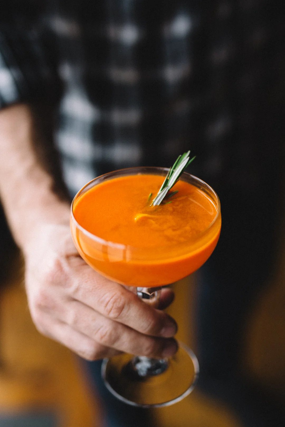 A person holding a stemmed glass of orange cocktail garnished with fresh herbs.