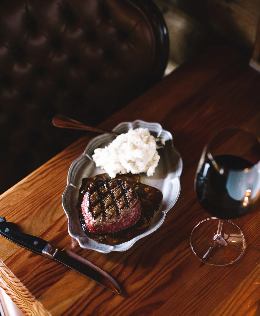 A plate with a grilled steak, mashed potatoes, and gravy, next to a glass of red wine on a wooden table.