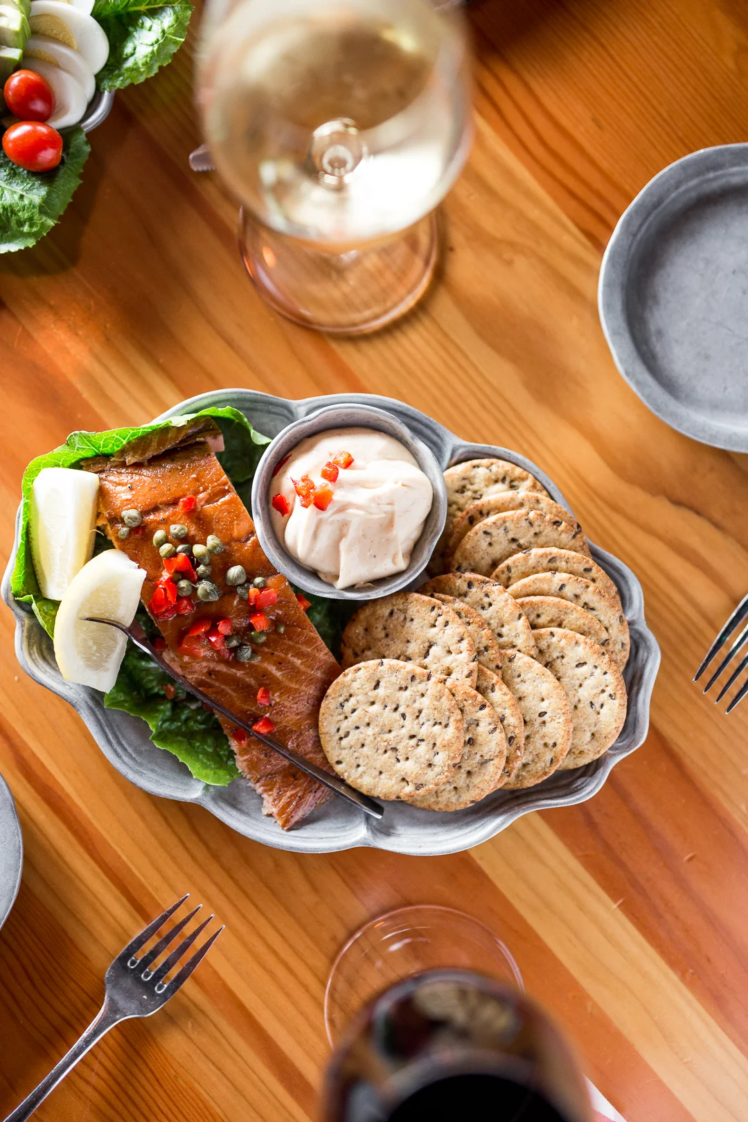 Plate of smoked salmon with lemon wedges, served with capers, a dollop of creamy sauce, and assorted crackers, on a wooden table with a glass of white wine and a fork nearby.