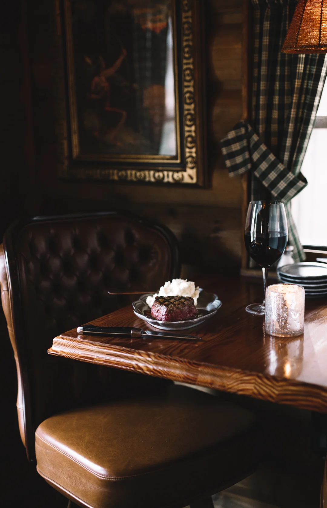 A rustic wooden table set for a meal with a plate holding a grilled steak with char marks, mashed potatoes, a glass of red wine, a lit candle, and a fork and knife. In the background, a framed painting of a dancer is on the wall, and a window with pl