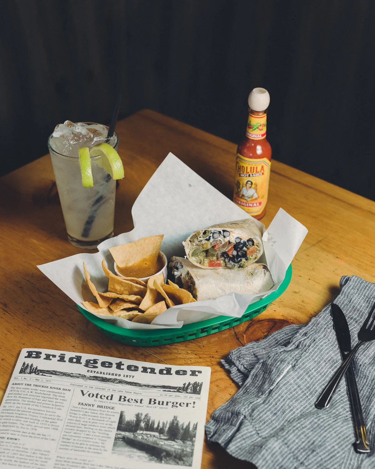 A basket with a burrito, tortilla chips, and salsa on a wooden table, accompanied by a drink with lime wedges and a bottle of hot sauce. A napkin, fork, and knife are also on the table, along with a newspaper.