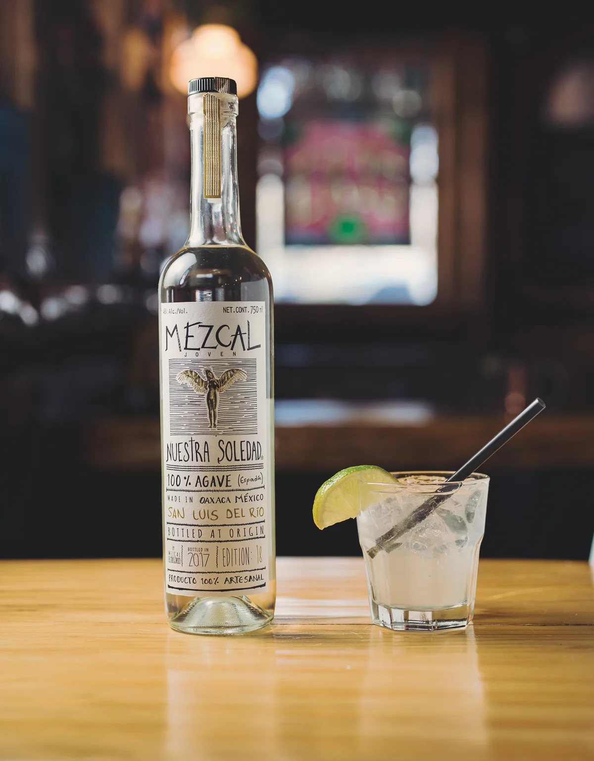 A bottle of mezcal and a glass of cocktail with lime wedge on a wooden table in a bar, with blurred background.