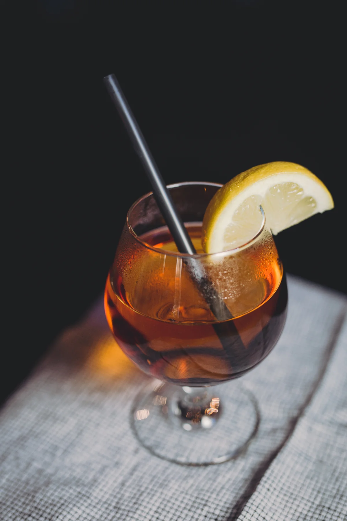 A cocktail glass filled with an amber-colored drink, garnished with a lemon wedge and a black straw, set on a white textured cloth surface against a dark background.