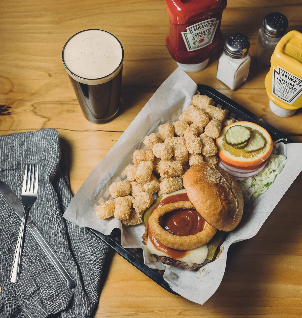 A cheeseburger with onion rings, pickles, and vegetables on a bun, served with fried chicken bites, sliced cucumber, tomato, onion, and lettuce, along with a glass of dark soda, ketchup, mustard, salt, and pepper on a wooden table.