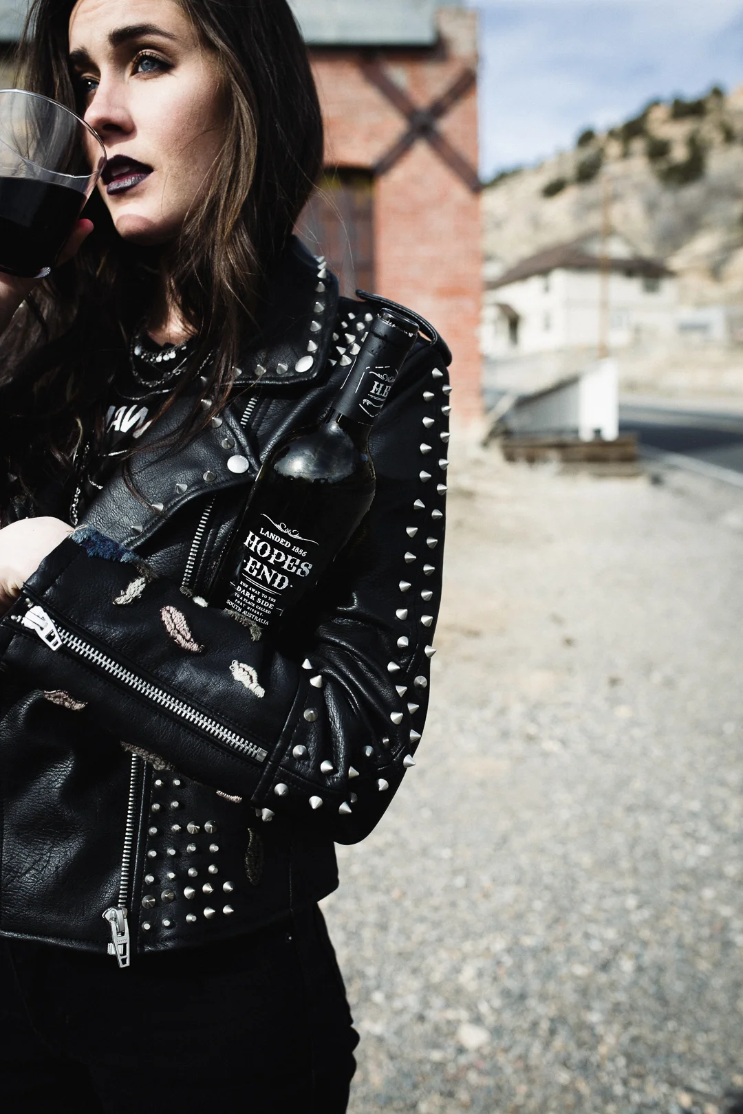 A woman with dark lipstick and long brown hair is wearing a black leather jacket with silver studs. She is holding a glass of dark beer or soda and has a bottle of beer with a label reading 'Hopes End' tucked into her jacket pocket. The background sh