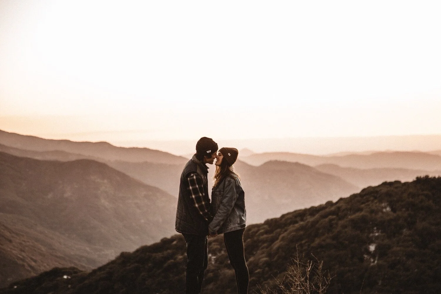 A couple standing close together on a mountain landscape at sunset, facing each other and holding hands, with layered mountains in the background.