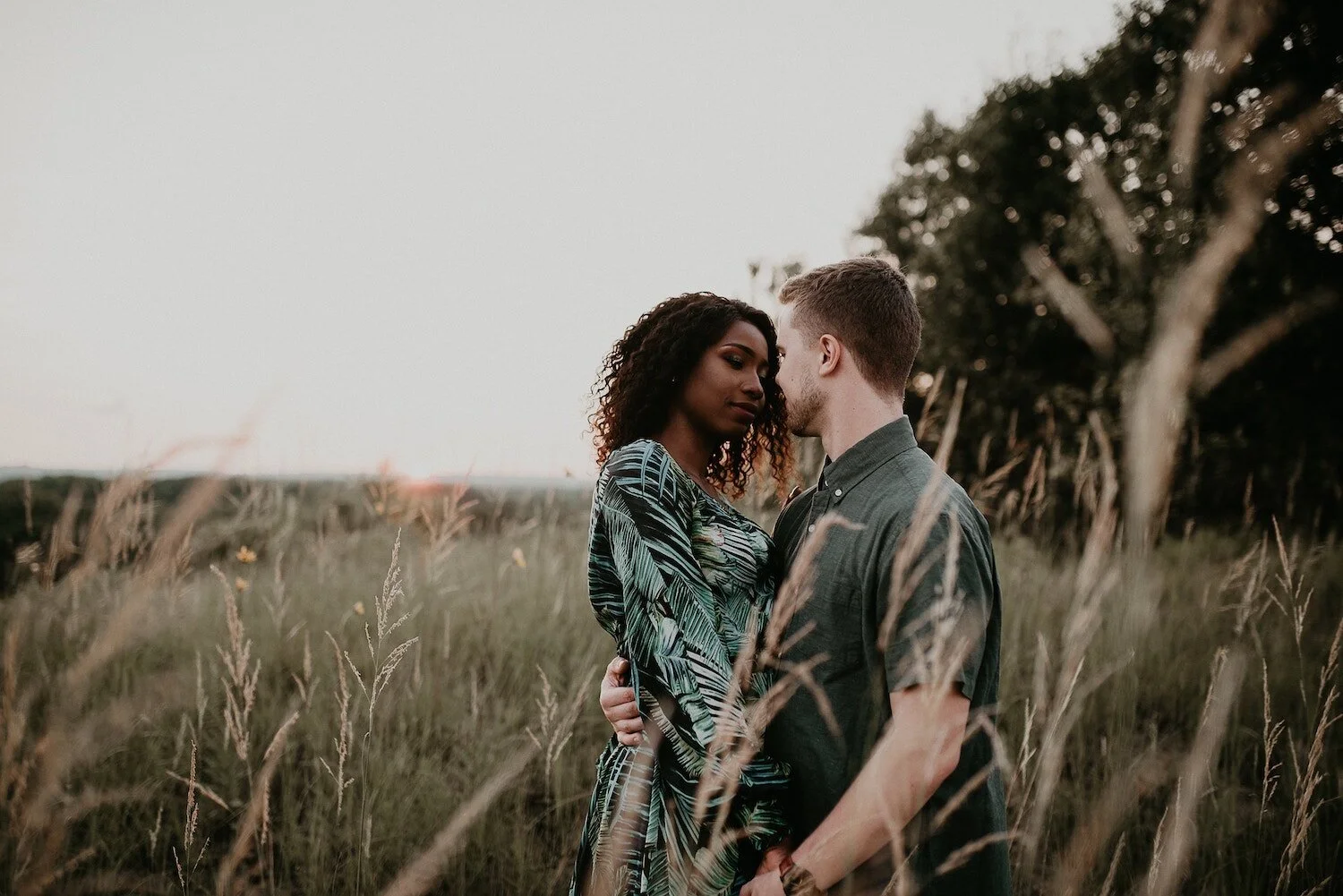 A couple standing close together in a grassy field at sunset, with trees in the background, embracing and looking into each other's eyes.