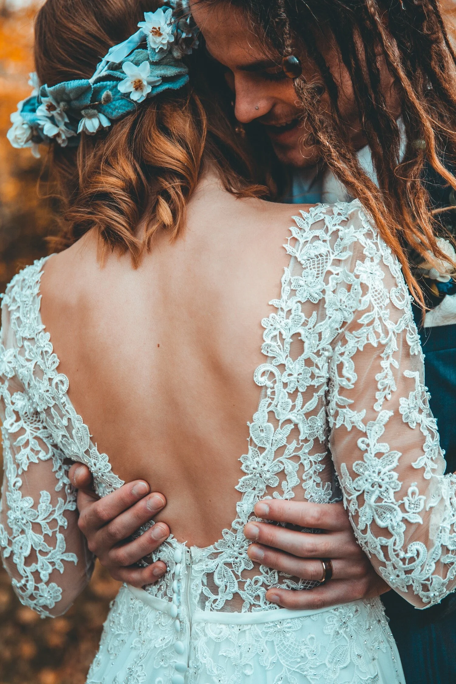 A couple embraces, with the woman wearing a white lace dress with an open back and floral embroidery, and the man holding her from behind with both hands on her waist. The woman has reddish hair with a blue floral headband. The background is blurred 