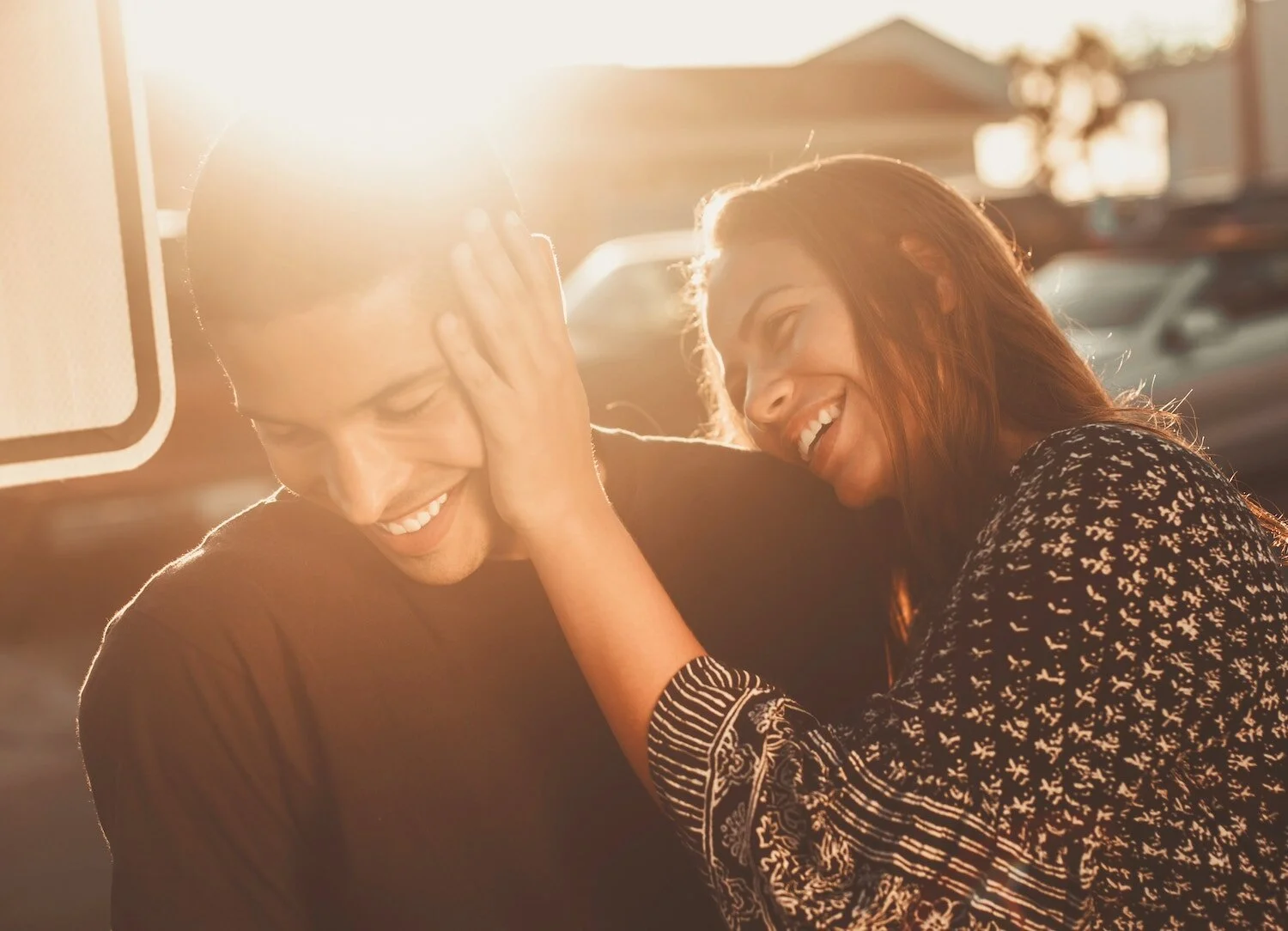 A man and woman laughing and sharing a joyful moment outdoors during sunset, with sunlight shining behind them.