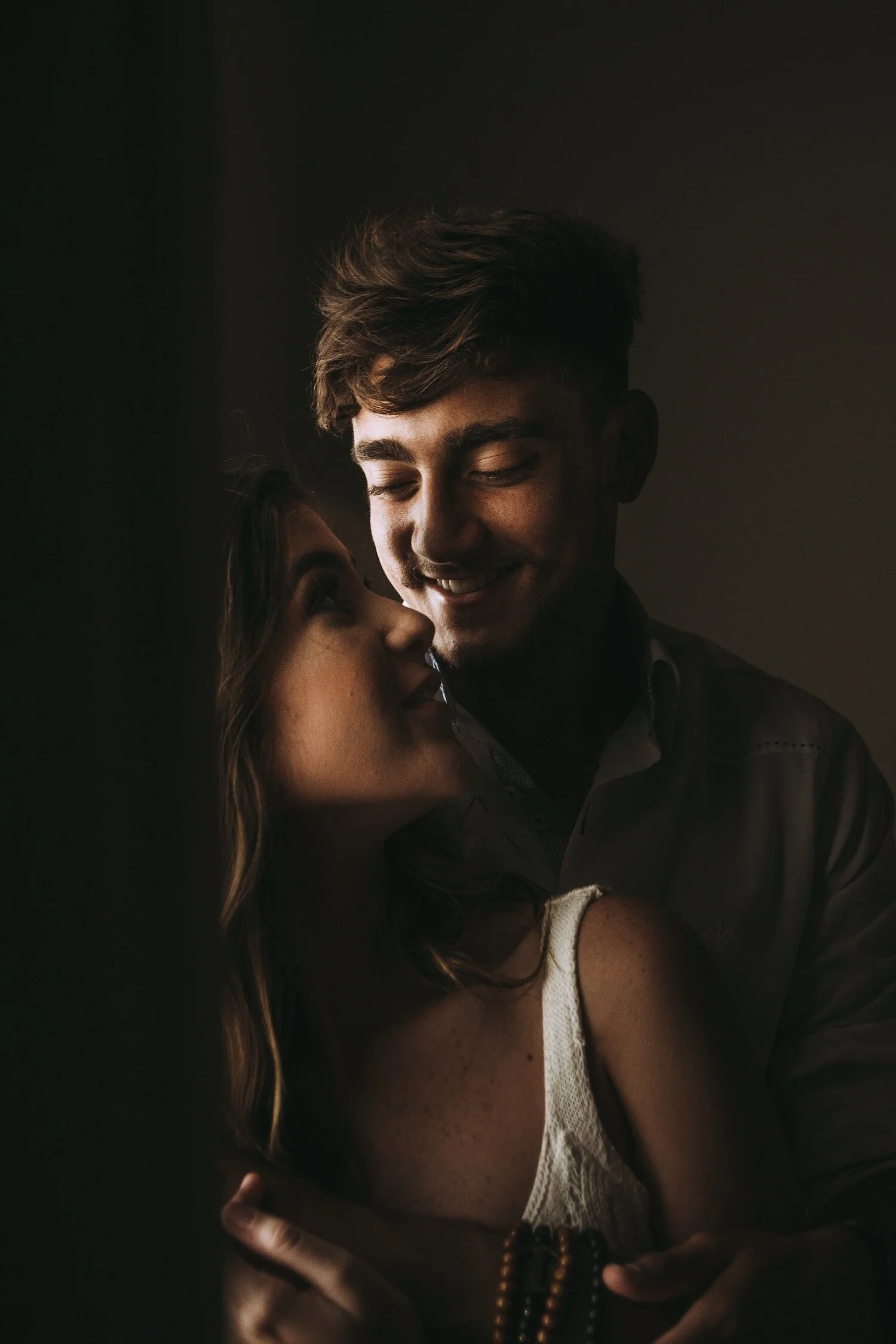 A young couple smiling and looking at each other in an intimate moment, with dim lighting creating a warm, romantic ambiance.
