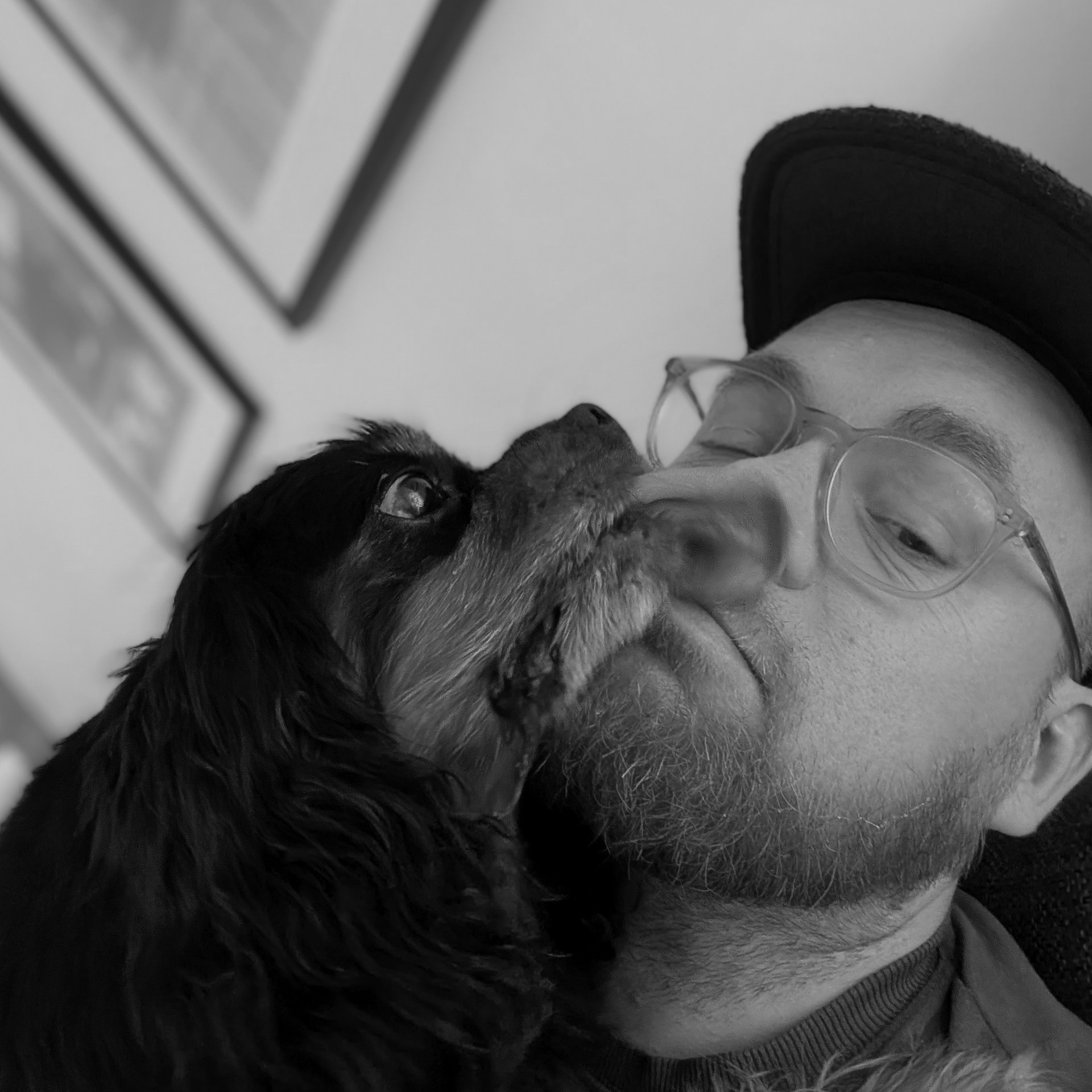 A person wearing glasses and a cap, lying down with a dog, with framed pictures on the wall in the background.