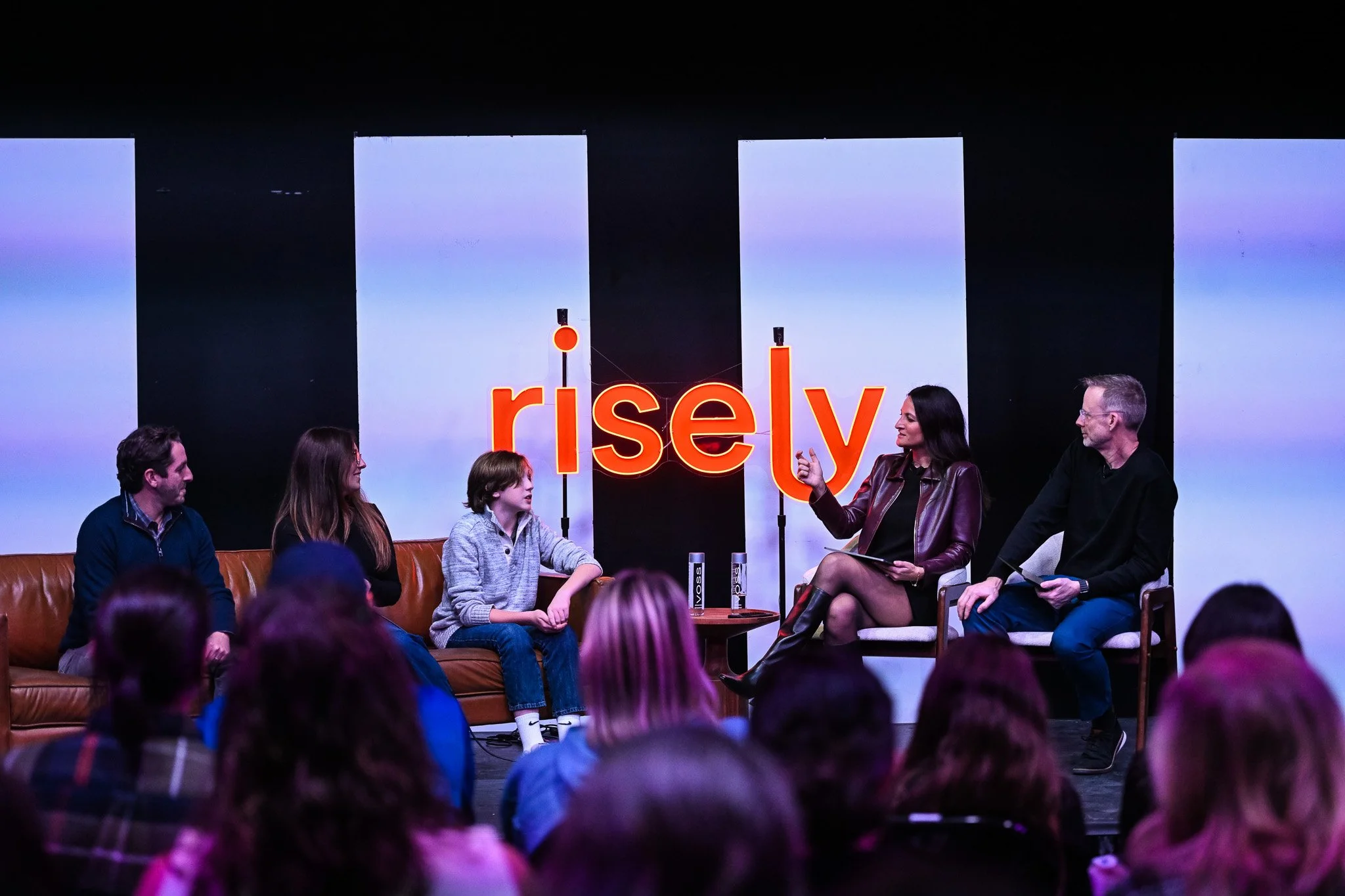 Panel discussion at an event called 'risely'. Five people are seated on stage, two women and three men, engaging with the audience. The woman on the right is speaking, and a young boy is sitting on a brown couch talking to a woman beside him. The aud