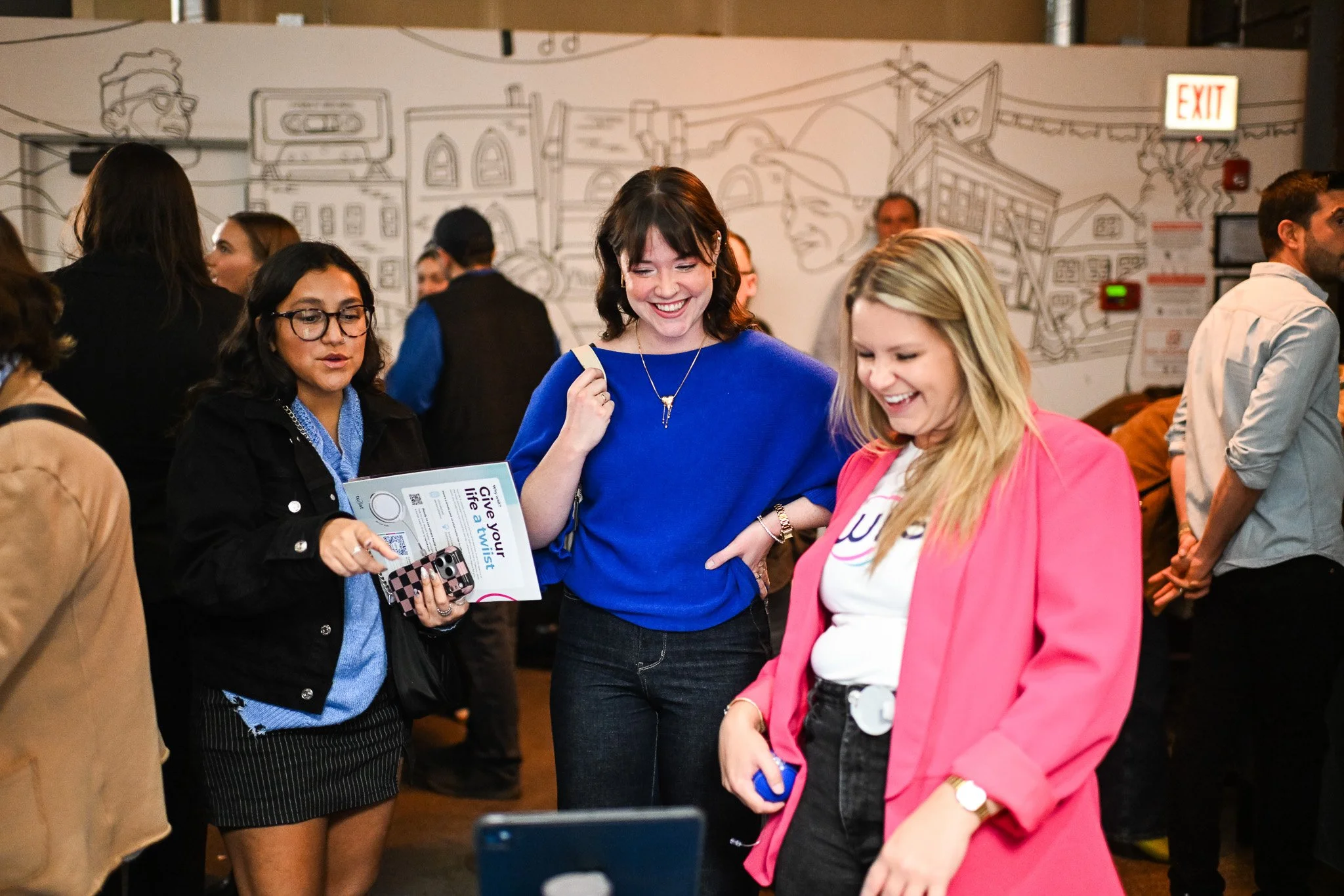 Group of diverse women smiling and talking at an indoor event, with a mural on the wall behind them.