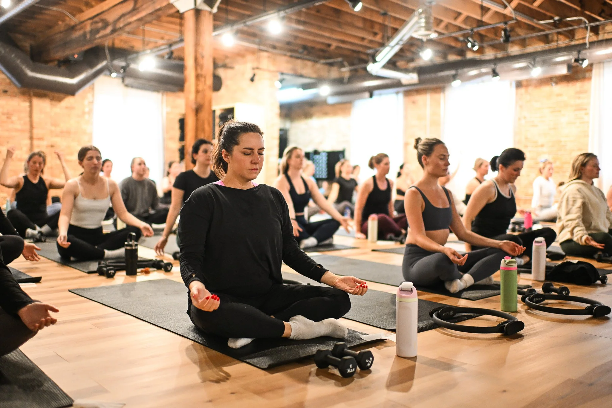 Group of women participating in a yoga class, seated on mats in a brick-walled studio, meditating and practicing breathing exercises.