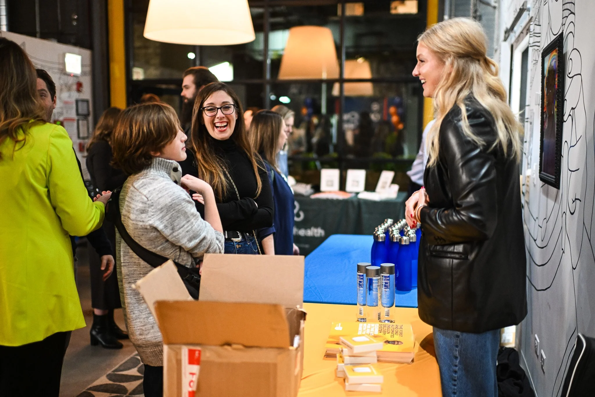 A group of people at a craft fair or event, talking to a woman with long blonde hair in a black leather jacket.  Books, water bottles, and other items are on a table.