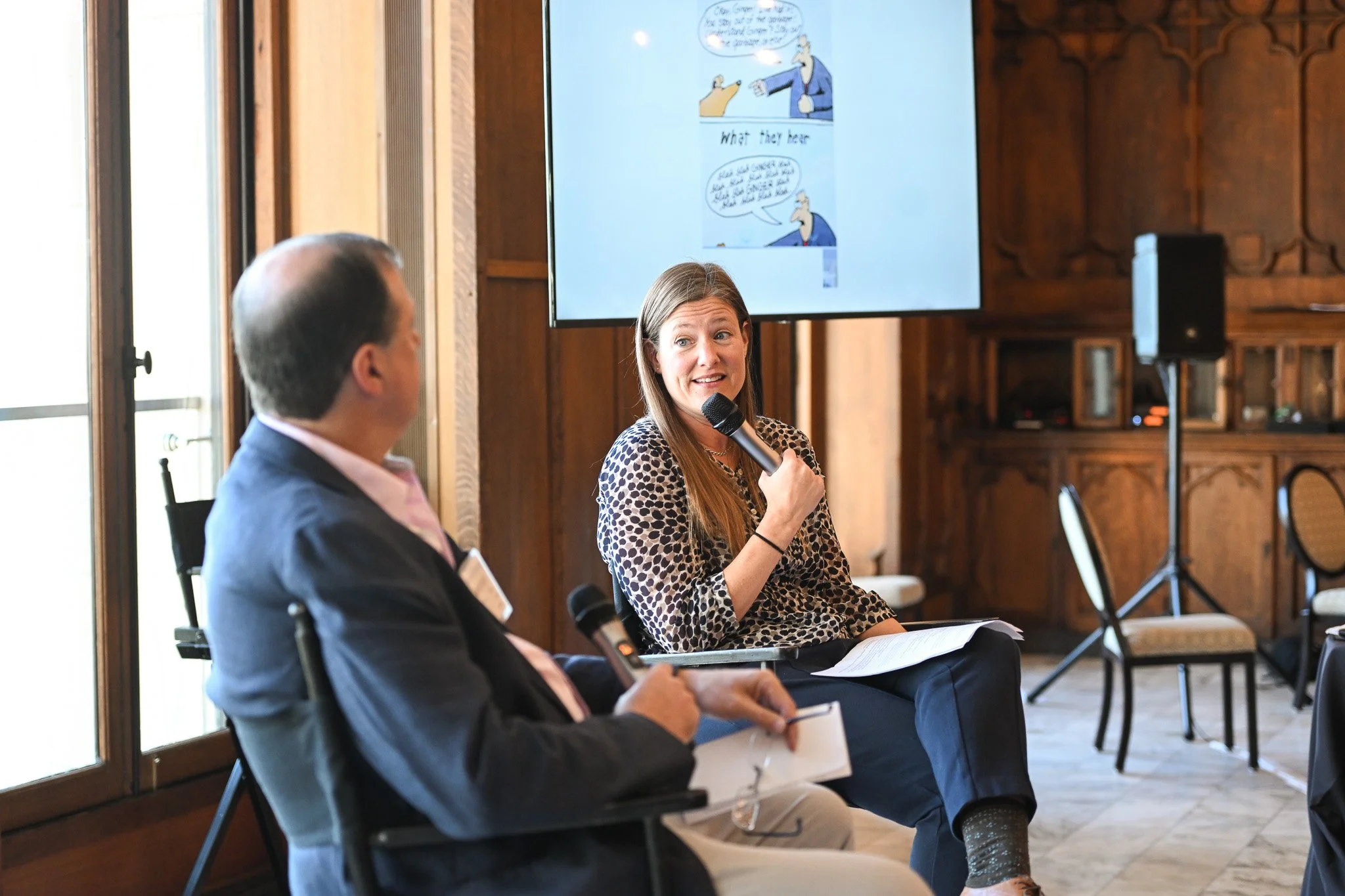 A woman speaking into a microphone during a panel discussion, with a man seated next to her holding notes, in a wood-paneled room with a large screen behind them displaying a cartoon comic strip.