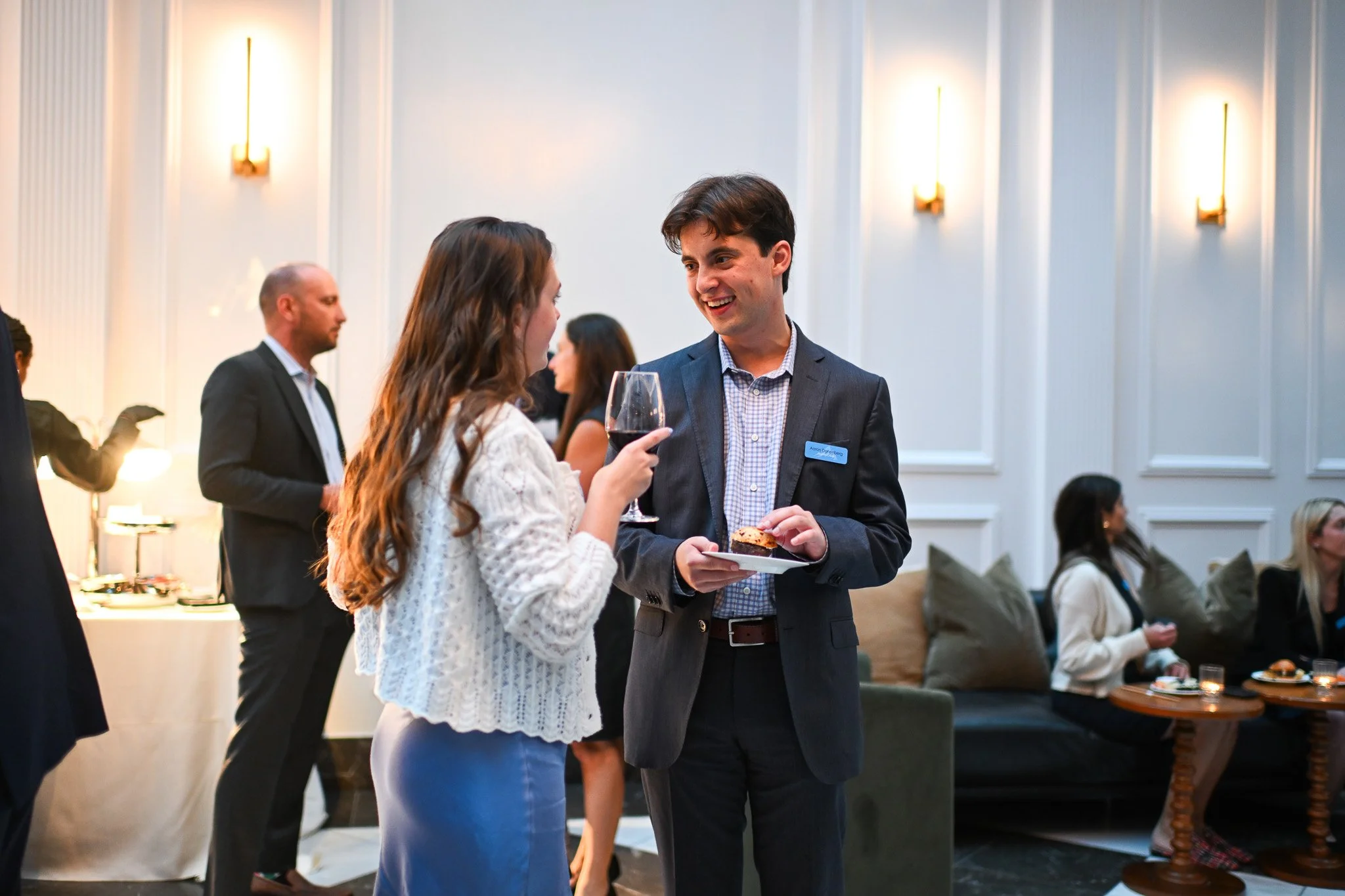 A man and woman are conversing at a social event, with the woman holding a glass of red wine and the man holding a plate with a canapé, in a well-lit, elegant room with other guests in the background.