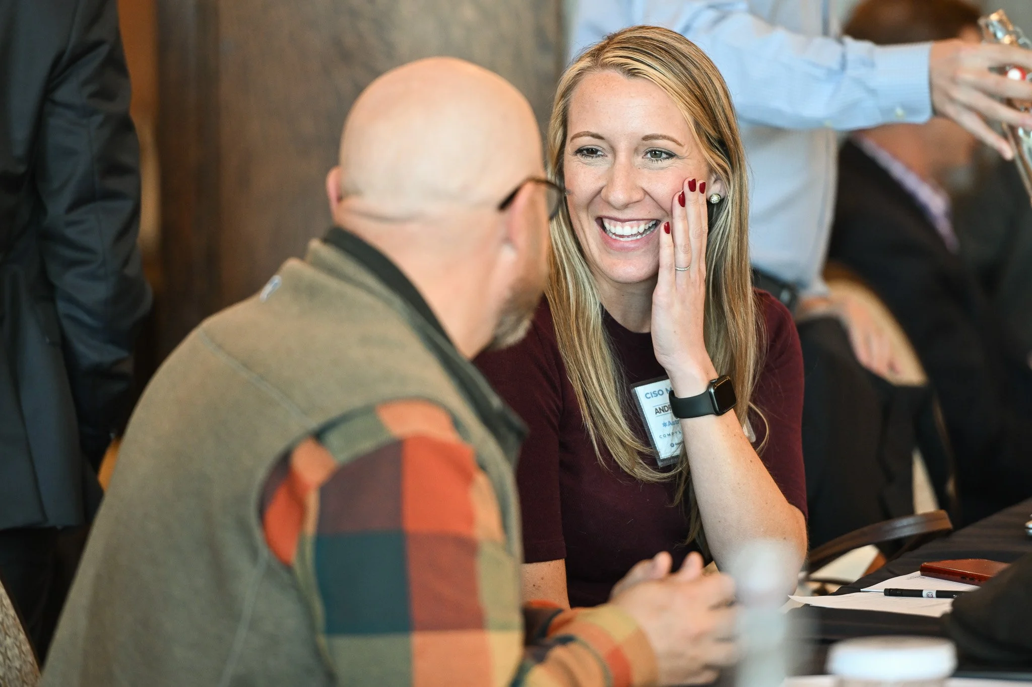 A woman with long blonde hair, wearing a maroon top, smiling and touching her cheek while engaging in conversation with a bald man in a plaid shirt in a conference or meeting setting.