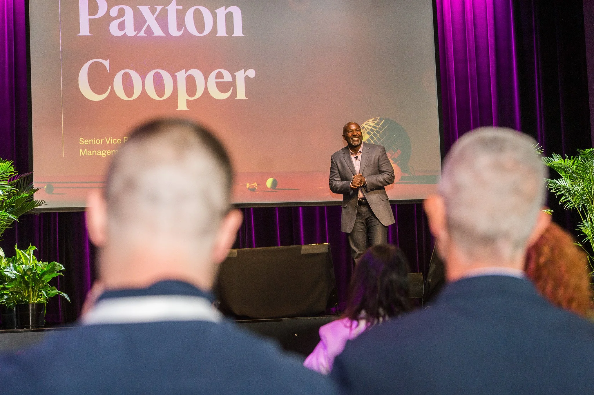 A man in a gray suit standing on a stage with purple curtains, smiling and speaking to an audience. The screen behind him displays the name Paxton Cooper and his title as Senior Vice President of Management.