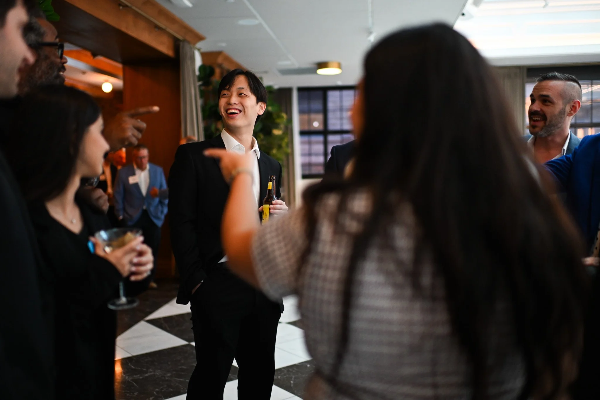 Group of people socializing and laughing at a party or gathering indoors, with a man holding a beer in the center.