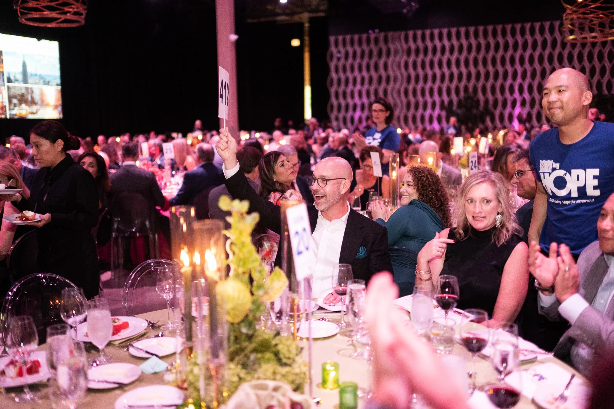 People seated at a banquet table in a large event hall with purple lighting. A man in a suit is smiling and raising a paddle with the number 20. Others are engaged in conversation, and waitstaff serve food. The table has floral centerpieces, wine gla