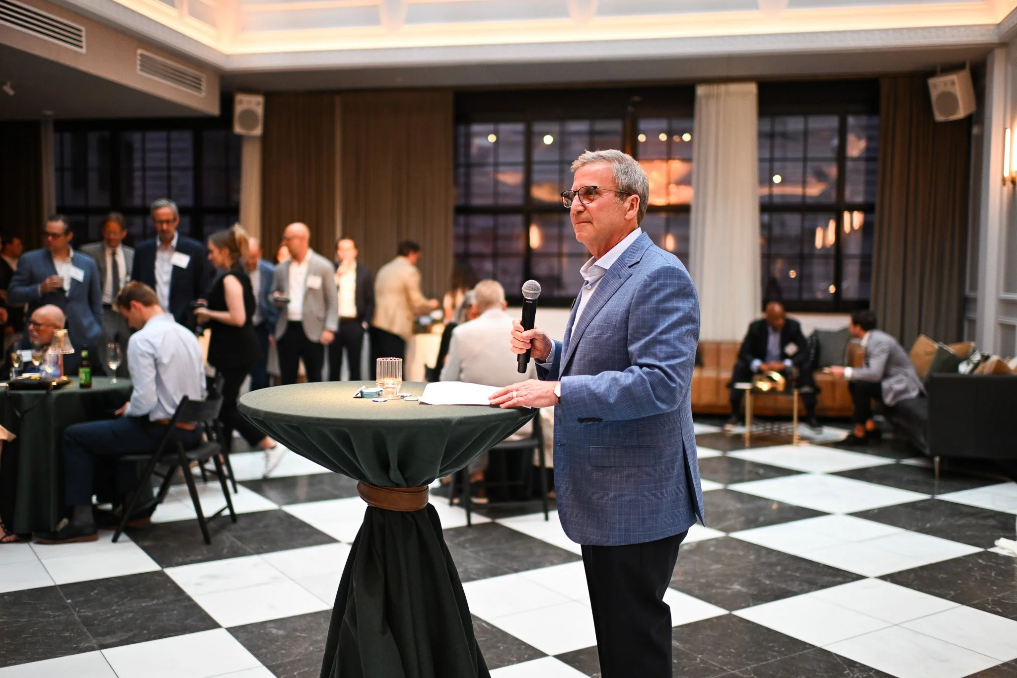 Man in suit speaking into a microphone at a standing table during a business conference or reception, with others socializing in the background.
