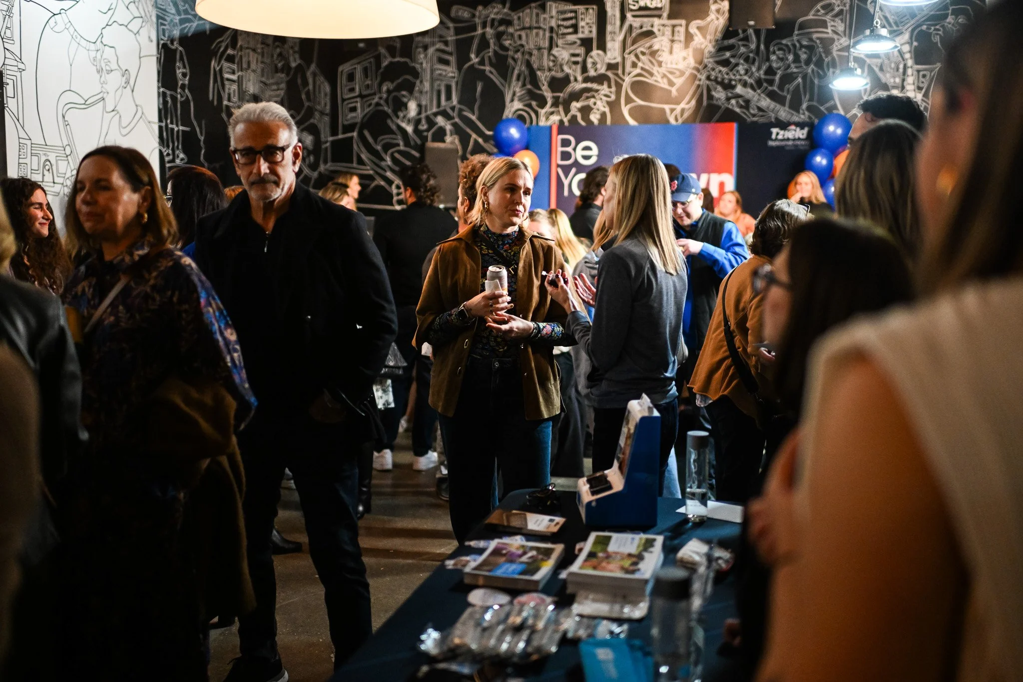 Crowd of people chatting at an indoor event with a black wall featuring white line art in the background and a sign that says 'Be Your' with balloons nearby.