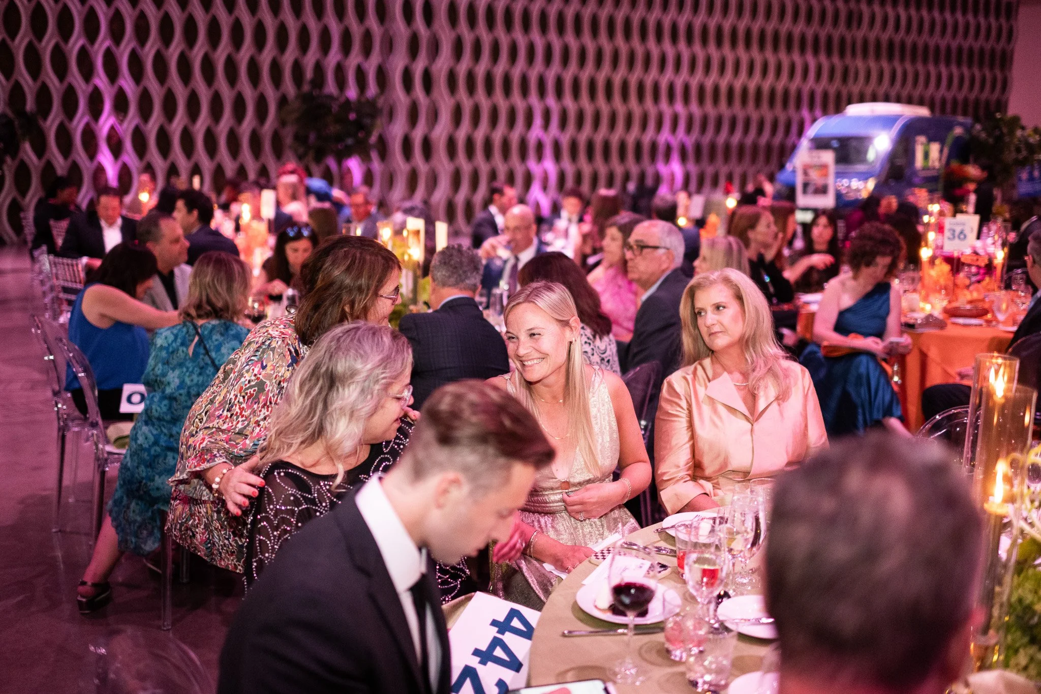 People seated around tables at an indoor banquet or gala dinner, engaging in conversations, with decorative lighting and a patterned wall in the background.
