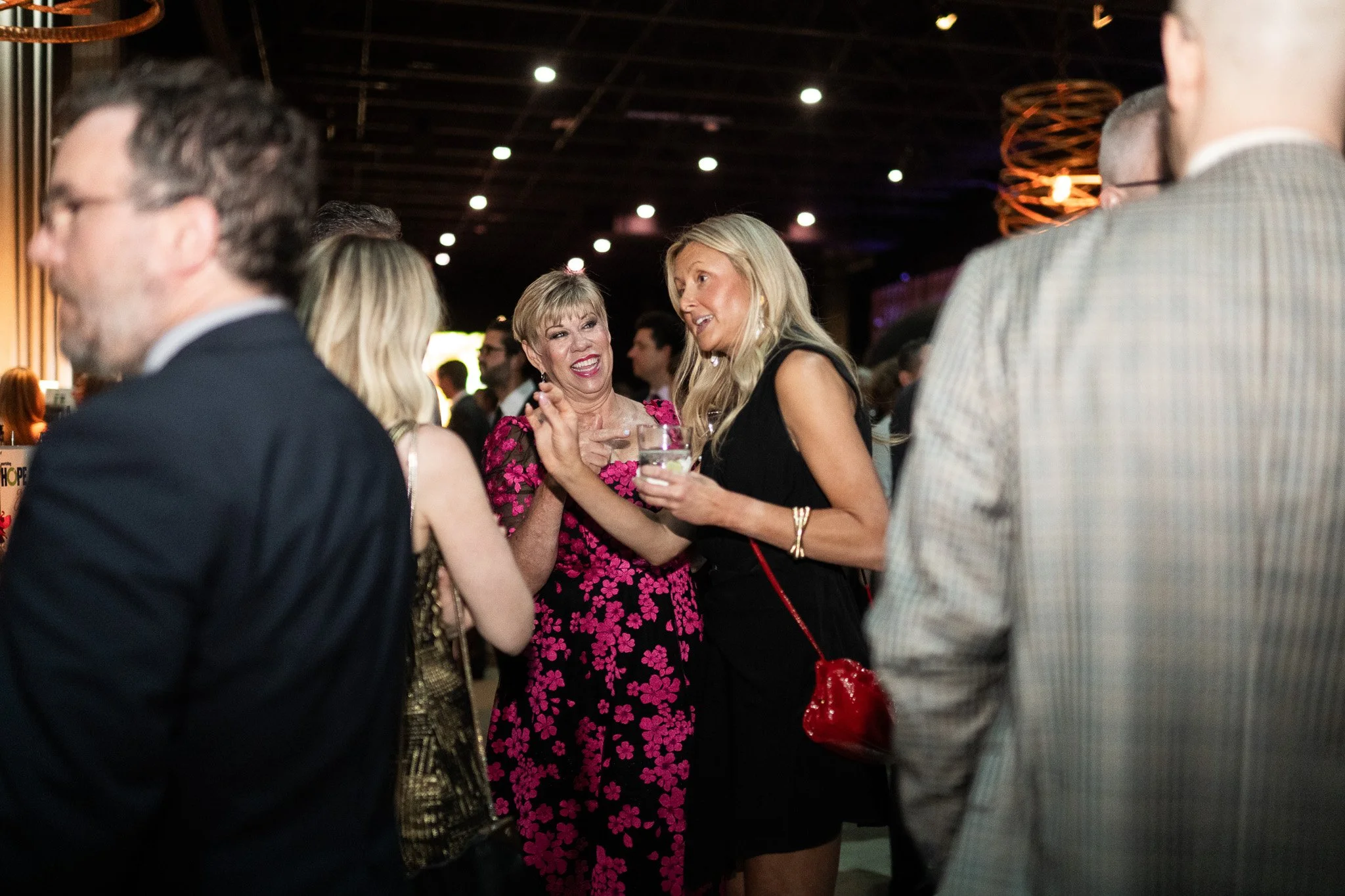 People socializing at an indoor event or party, with two women in conversation at the center, one holding a drink.