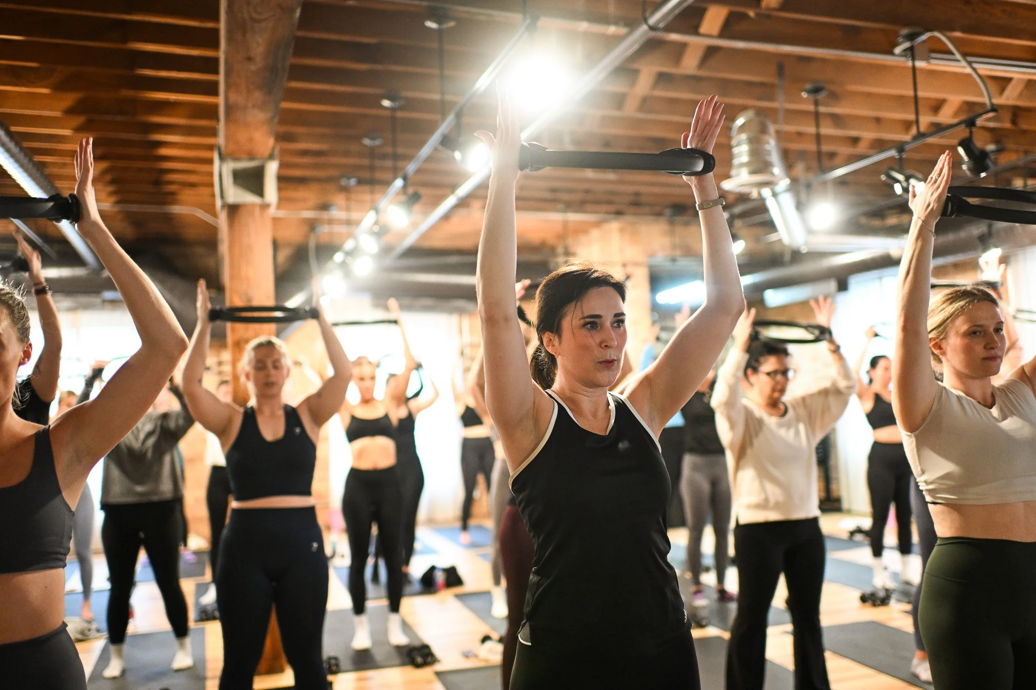 Women participating in a fitness class doing overhead exercises with suspension trainers in a wood-paneled studio.