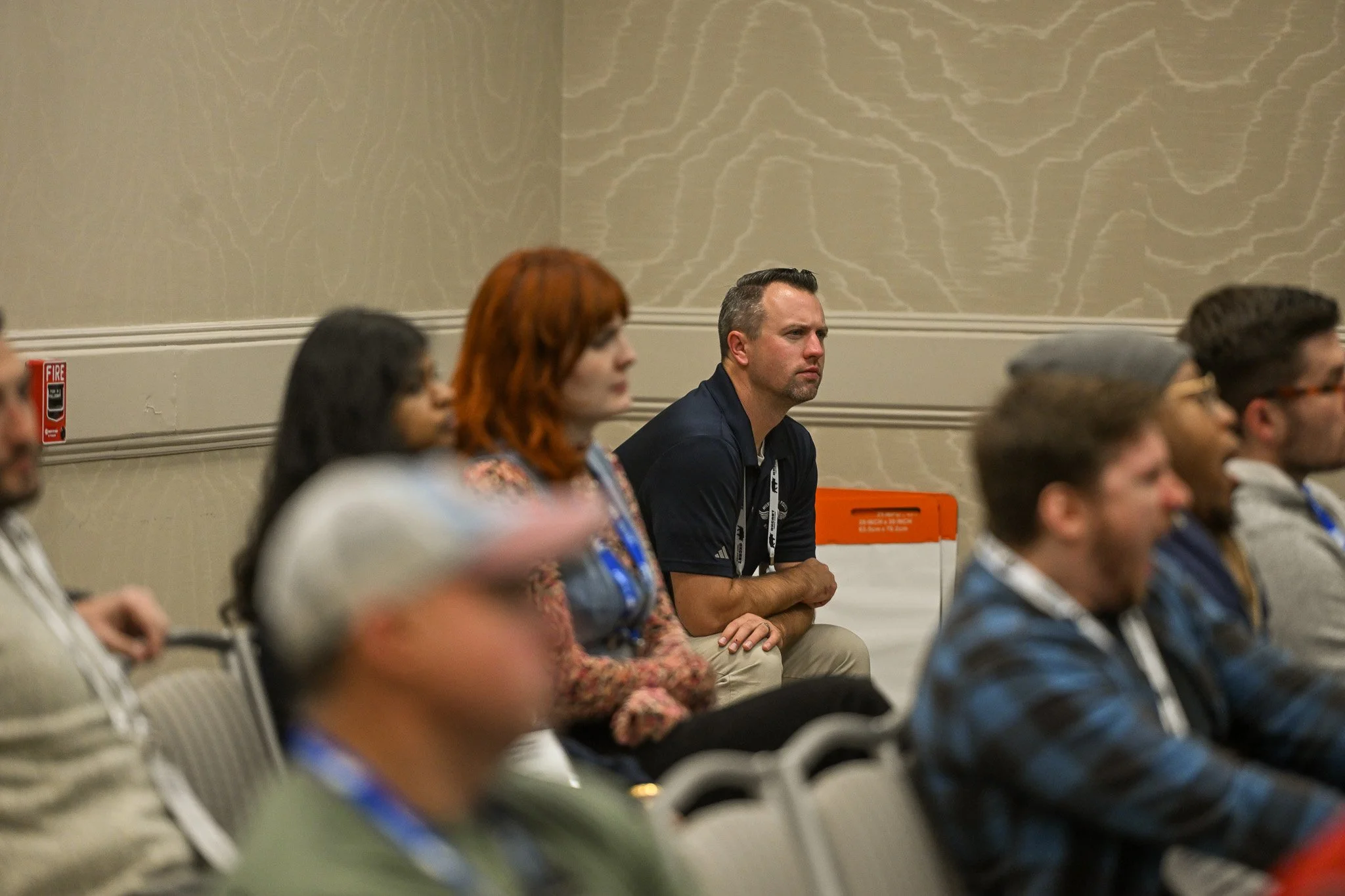 A group of people attending a conference or seminar, sitting in rows of chairs in a conference room, some wearing lanyards, focused on a presentation or speaker.