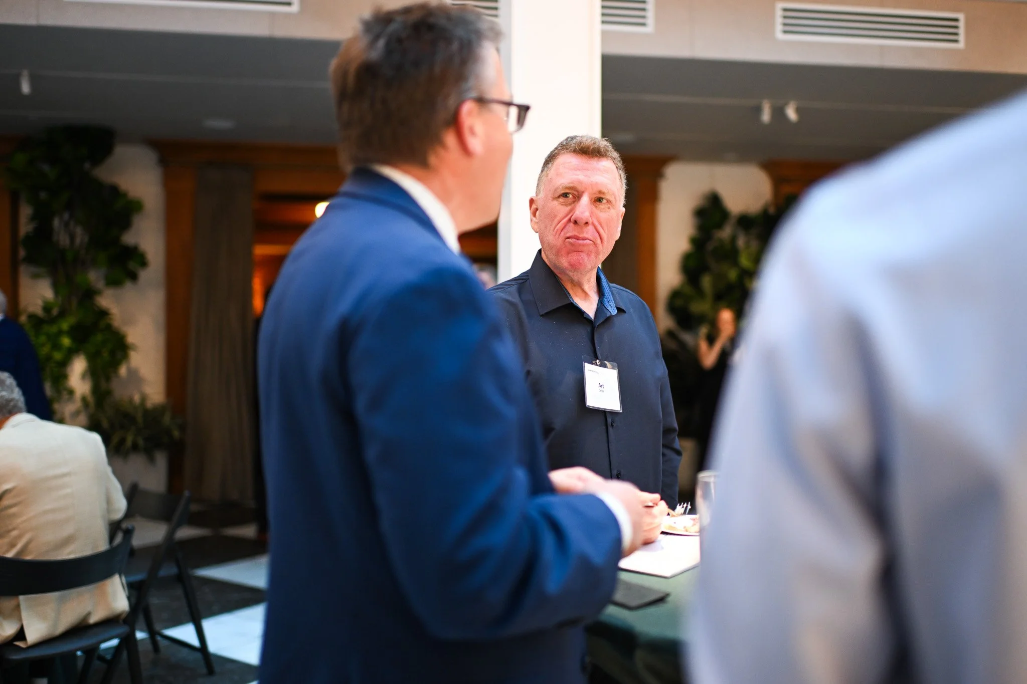Three men engaged in conversation at a social event, with one man in focus wearing a dark shirt and a name tag, while another man in a blue suit is in the foreground and a third person with a light-colored shirt is partially visible on the right. Peo