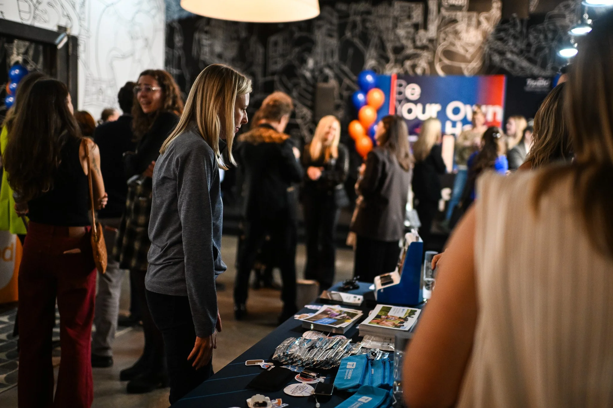 A group of women at an indoor event, with one woman in the foreground looking at items on a table, including magazines and promotional materials, while others chat and mingle in the background, with balloons and a sign that says 'Be Our Own Hero'.