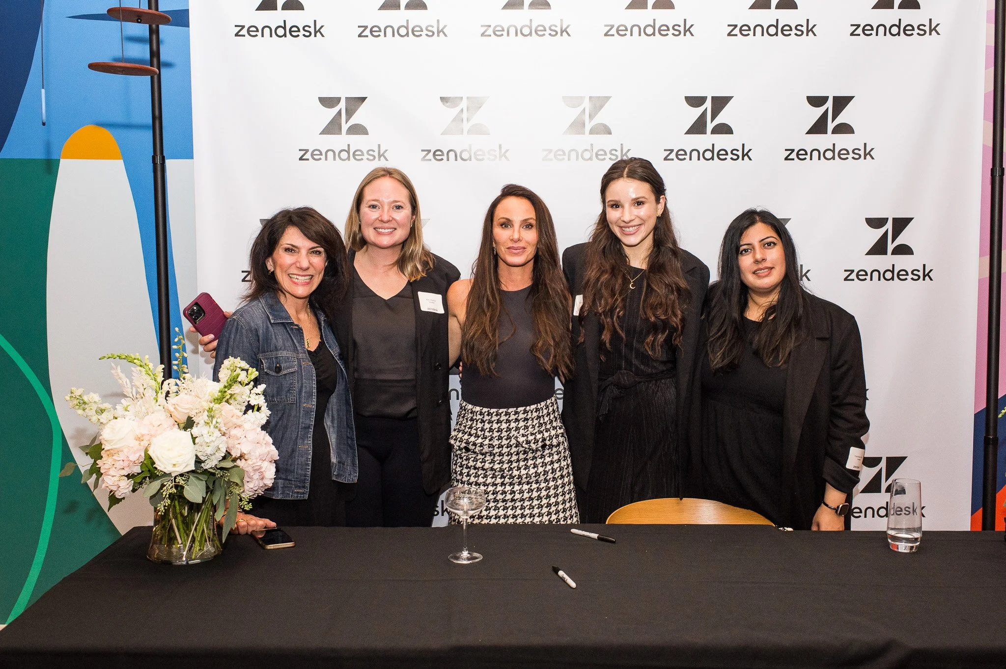 Five women standing together at a Zendesk event, smiling, with a white backdrop featuring the Zendesk logo behind them. There is a table in front of them with a floral arrangement, two markers, and a glass of water.