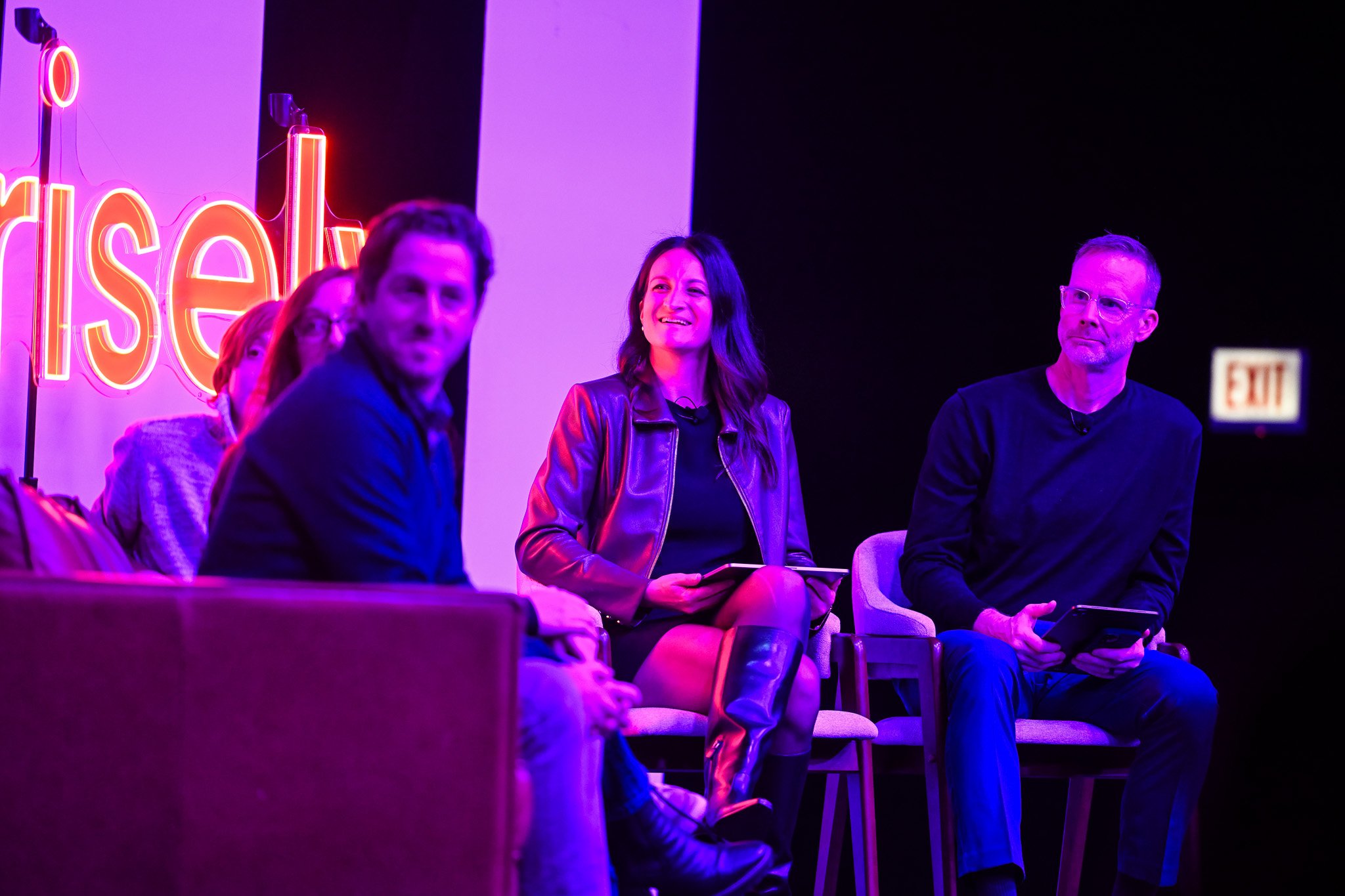 People sitting on stage at a panel discussion, illuminated with purple and pink lighting, with a partially visible neon sign in the background.