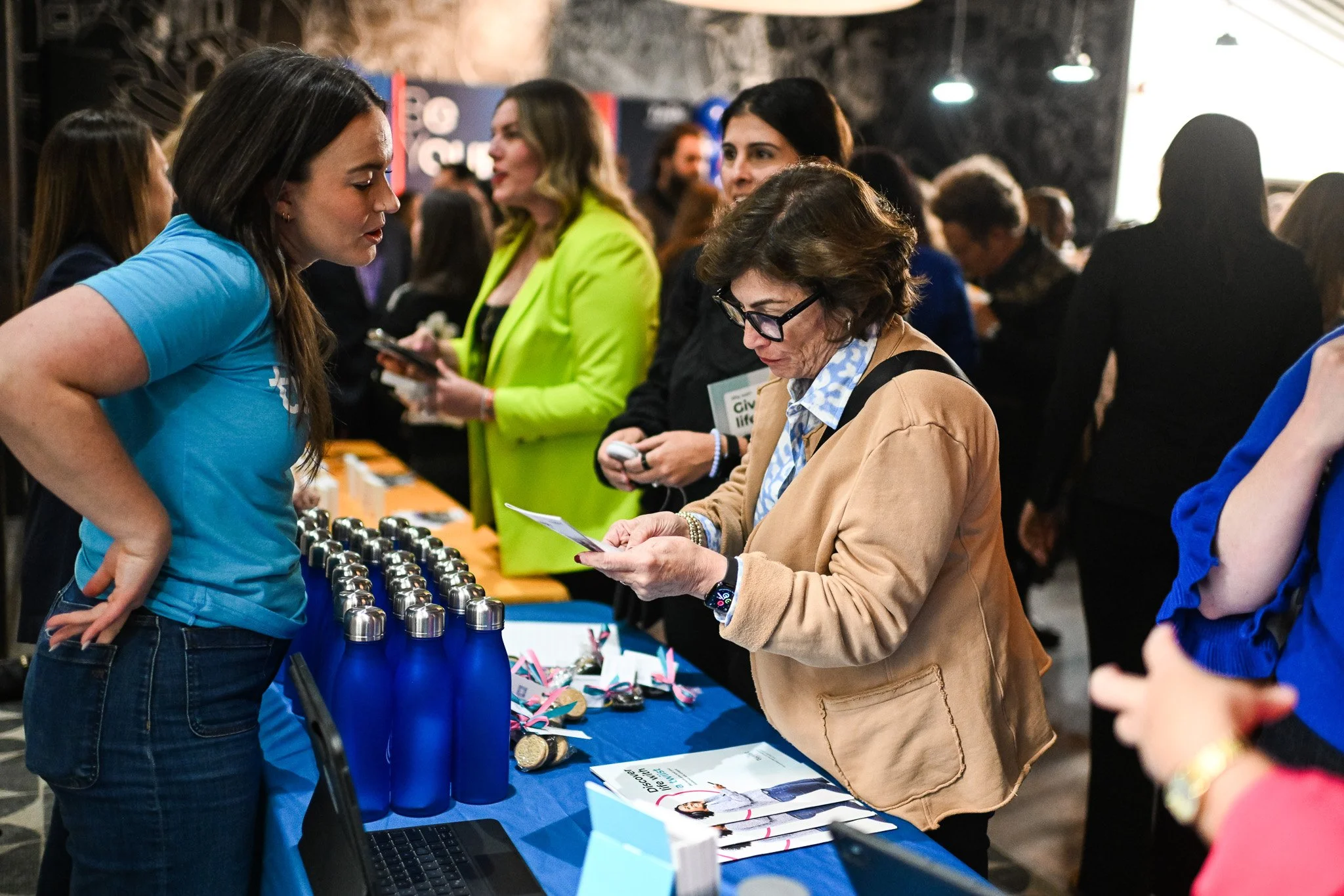 Women engaged in conversation at a booth with blue water bottles and brochures during an indoor event.