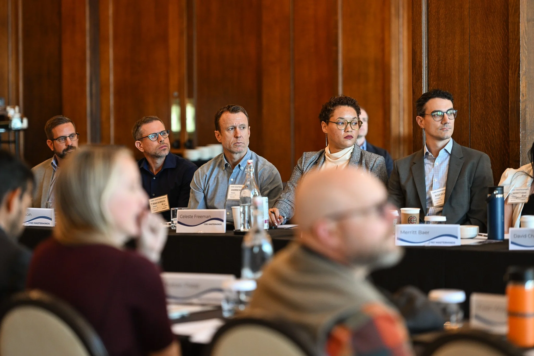 Attendees sitting at a conference table in a wood-paneled room, listening to a presentation. The table has name tags, water bottles, and cups.