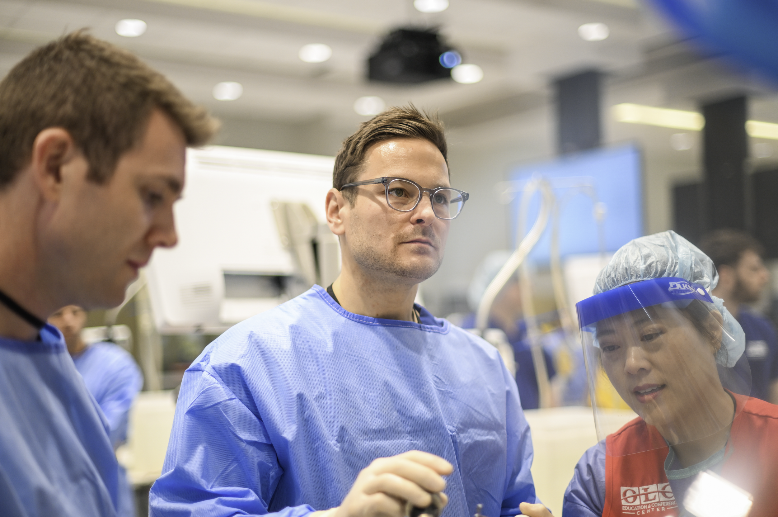 Medical professionals in scrubs and protective gear in a hospital setting, focused on a procedure or discussion.