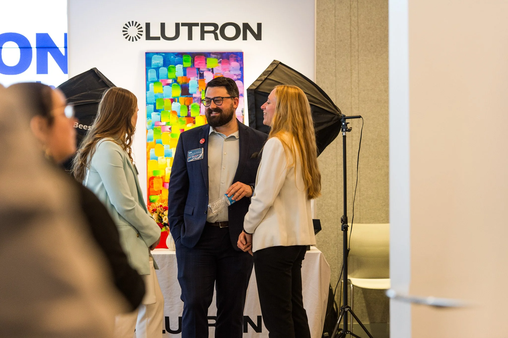 People talking at an event with a Lutron sign, colorful abstract artwork, and professional lighting equipment in the background.